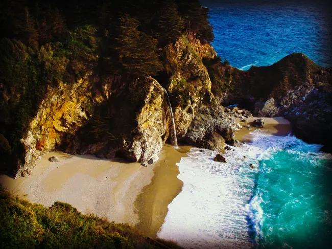 Aerial view of a coastal cliff with a waterfall onto a sandy beach and turquoise ocean, green vegetation on the cliffs